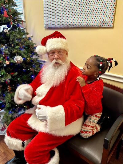 A young girl dressed in holiday clothes looks over Santa's shoulder during a precious game of peek-a-boo during his visit to Tidelands Health Center for Pediatric Development in Georgetown.